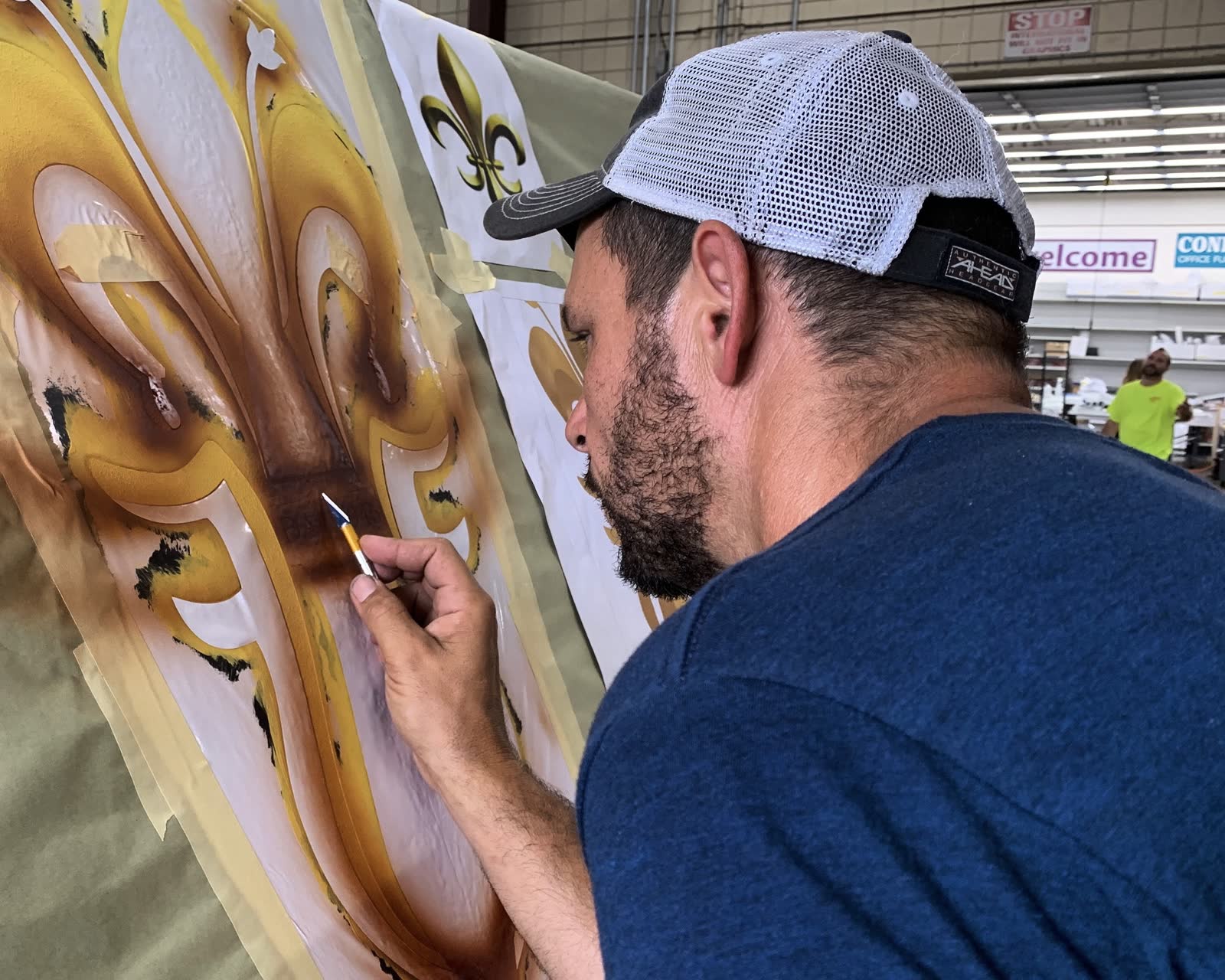Artist airbrushing a large gold fleur-de-lis painting on a panel in a shop