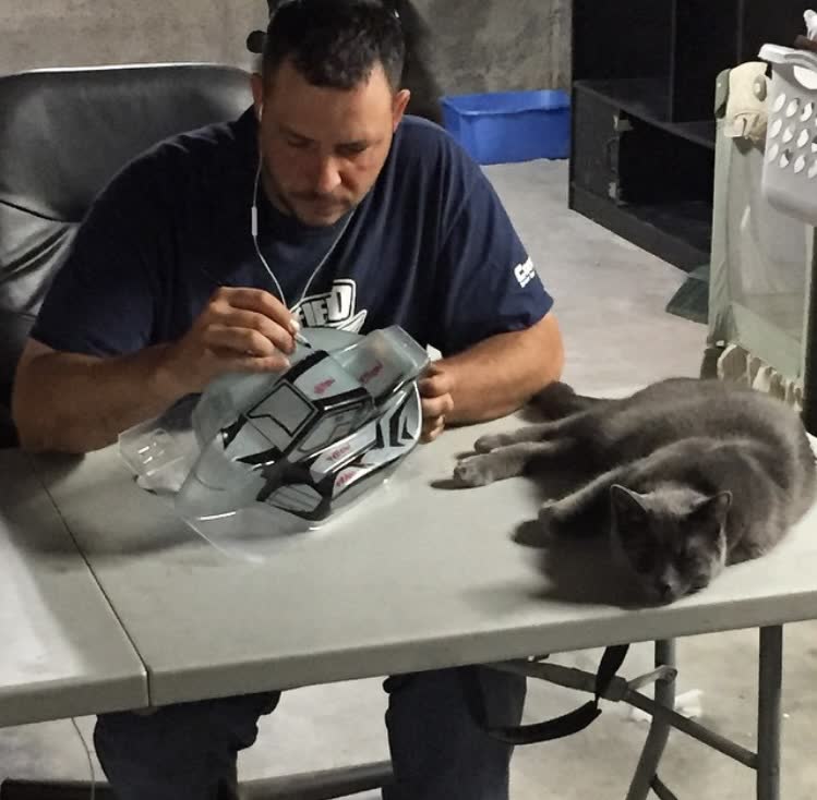 Artist masking a clear Lexan RC body at a worktable with a gray cat nearby